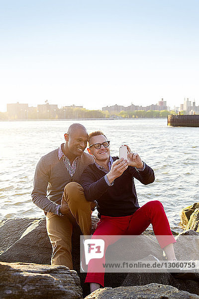 Happy boyfriends taking selfie while sitting on rocks against river on sunny day