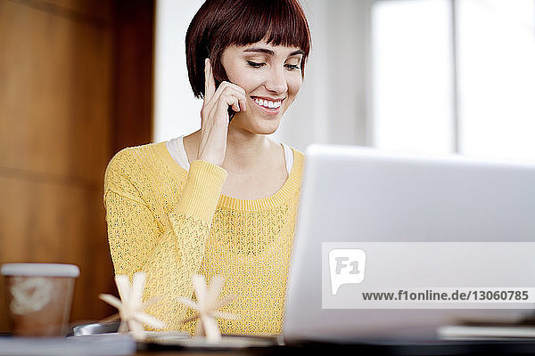 Glückliche Geschäftsfrau telefoniert  während sie im Büro einen Laptop benutzt