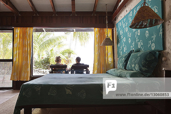 Rear view of couple relaxing on lounge chairs seen through bedroom window