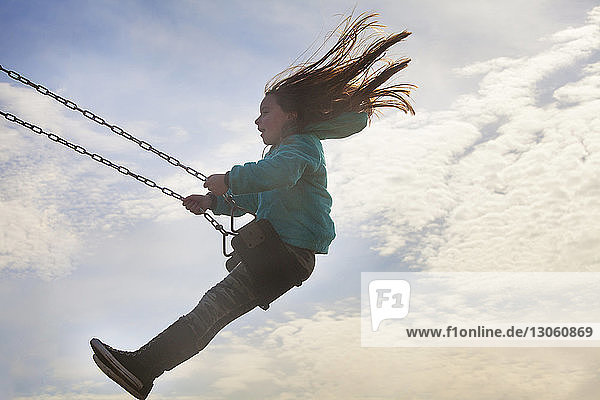 Side view of girl swinging against cloudy sky
