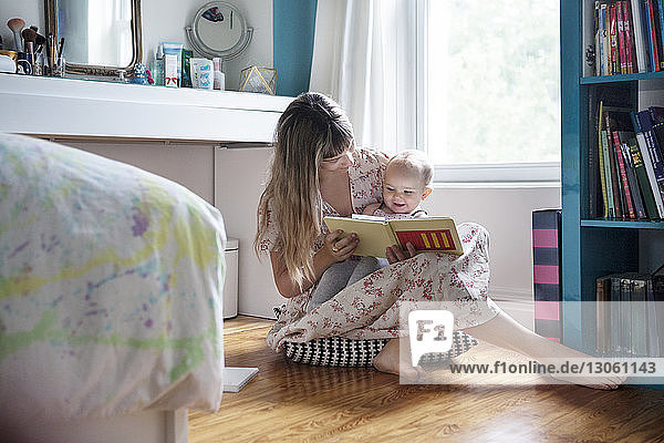 Mother reading book with cheerful baby while sitting on floor