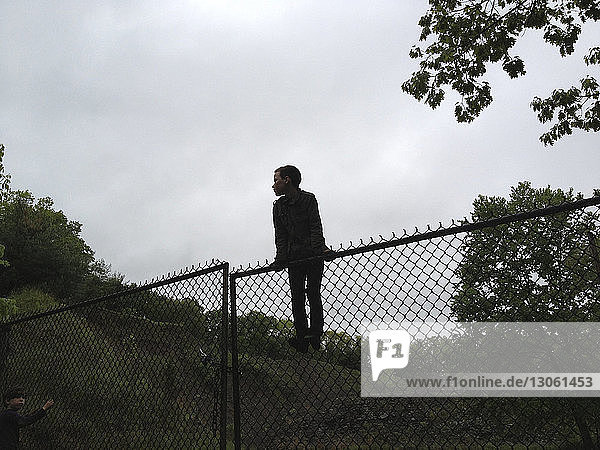 Junge in voller Länge auf Kettengliedzaun stehend auf Feld gegen Himmel
