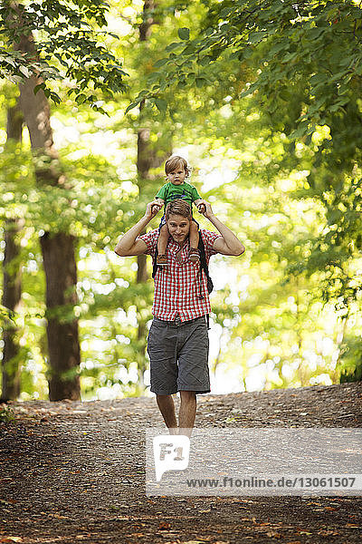 Porträt eines Vaters  der seinen Sohn auf den Schultern trägt  während er im Wald auf einem Feldweg geht