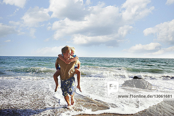 Vater gibt seinem Sohn Huckepack  während er an einem sonnigen Tag am Meeresufer spazieren geht