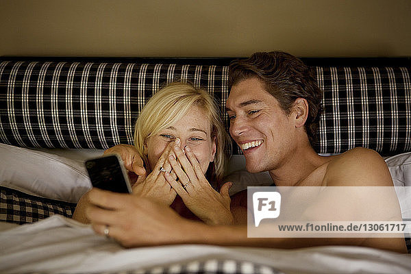 Glückliches Paar  das zu Hause im Bett liegend auf sein Handy schaut