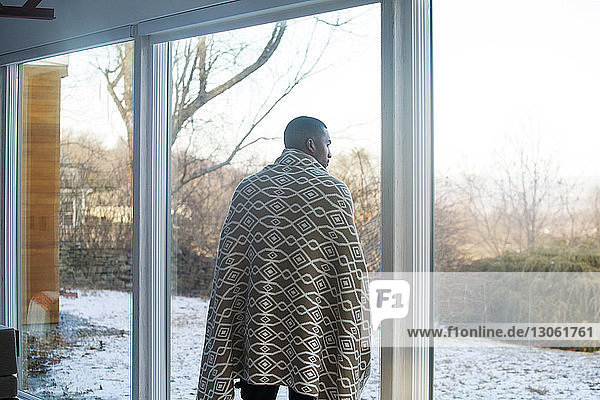 Rückansicht eines mit einer Decke bedeckten Mannes  der zu Hause am Fenster steht