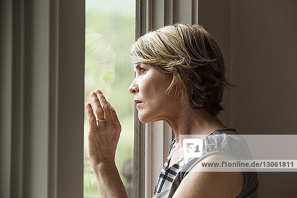 Seitenansicht einer Frau  die zu Hause durch ein Fenster schaut