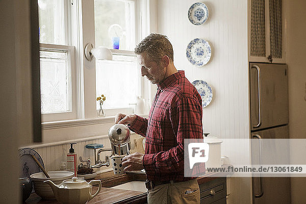 Seitenansicht eines Mannes  der Kaffee in einen Kaffeebecher gießt  während er zu Hause an der Küchentheke steht