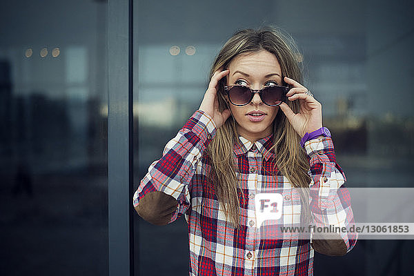 Beautiful woman holding sunglasses while looking away against building