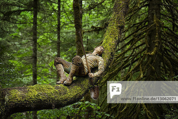Landwirt entspannt sich auf Baumstamm  während er im Wald eine Schaufel hält