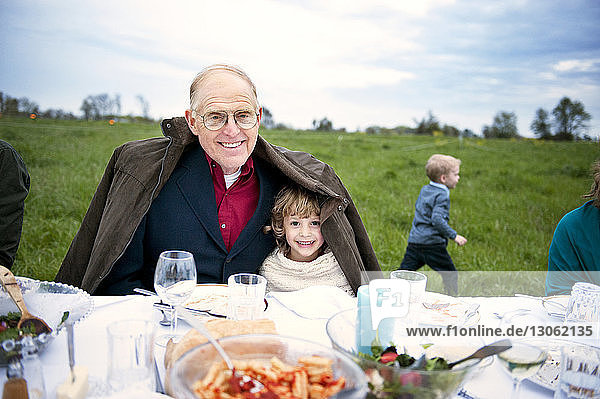 Portrait of happy girl sitting with grandfather at table on field