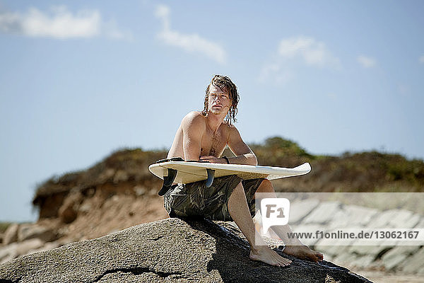 Man with surfboard looking away while sitting on rock