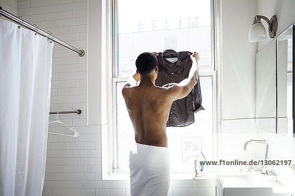 Rear view of man holding ironed shirt by window in bathroom