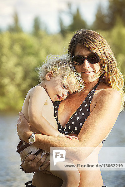Close-up of mother wearing swimwear carrying smiling son against lake