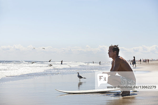 Thoughtful man with surfboard crouching at beach