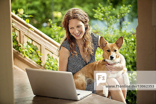 Woman using laptop computer while sitting with dog on porch