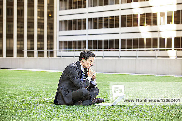 Nachdenklicher Geschäftsmann schaut auf Laptop-Computer  während er auf Grasfeld sitzt
