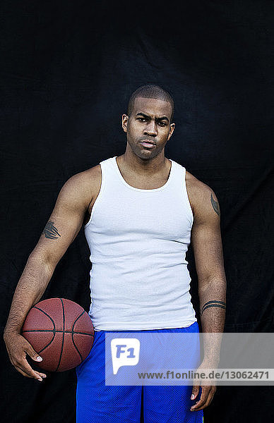 Porträt eines selbstbewussten Mannes  der Basketball hält  während er gegen eine schwarze Wand steht