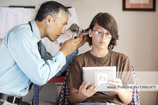Junge hält Tablett-Computer in der Hand  während der Arzt sein Ohr zu Hause untersucht