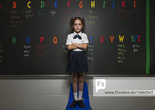 Portrait of confident schoolgirl standing on chair against blackboard