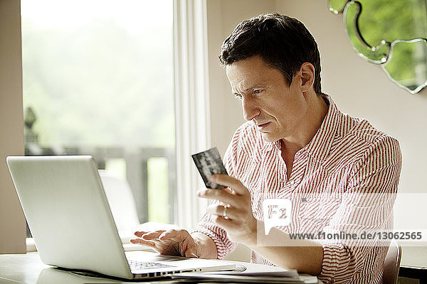 Mann hält Kreditkarte  während er zu Hause einen Laptop benutzt