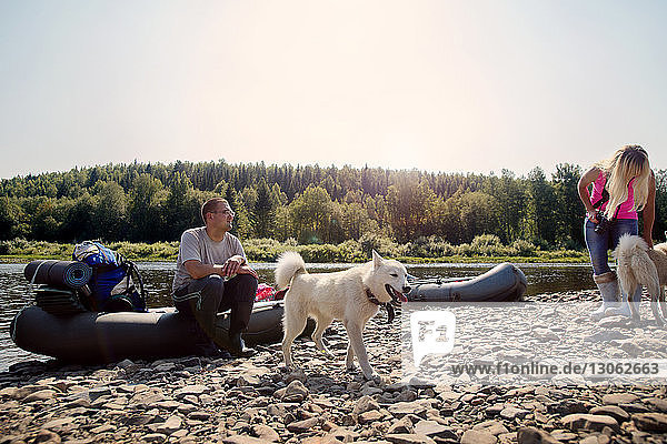 Freunde mit Hunden mit aufblasbaren Flößen am Flussufer bei klarem Himmel