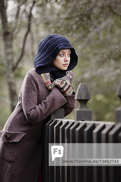 Woman in warm clothing looking away while leaning on fence