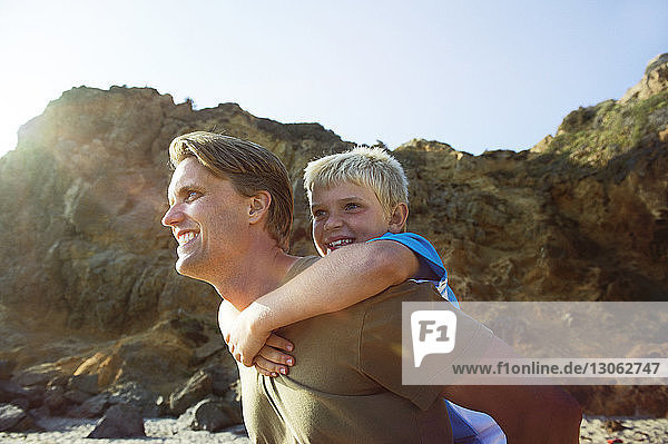 Happy father giving piggyback to son at beach against rock formation