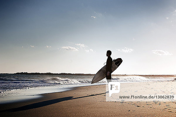 Seitenansicht eines Surfers  der ein Surfbrett trägt  während er am Strand am Ufer steht
