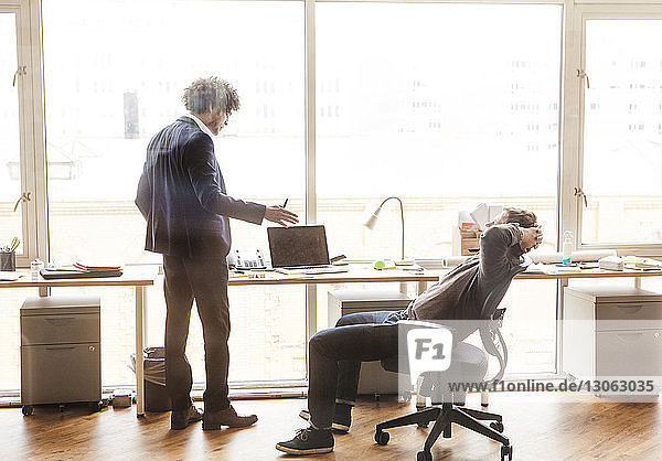 Geschäftsmann erklärt  während ein Kollege sich auf einem Stuhl im Büro entspannt