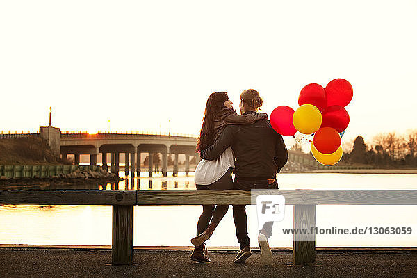 Rear view of couple with balloons sitting on railing during sunset