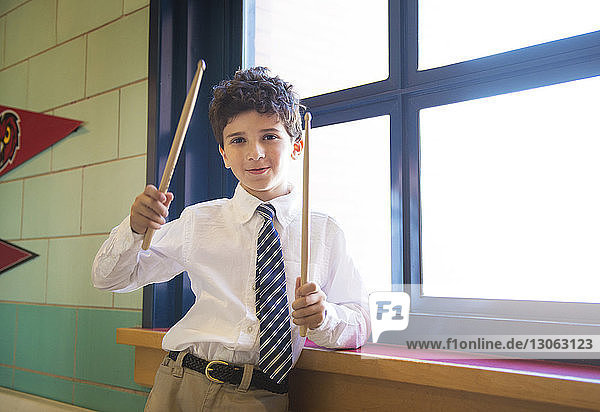 Portrait of schoolboy holding drumsticks while standing at window