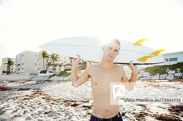 Porträt eines Mannes ohne Hemd  der ein Surfbrett trägt  während er am Strand vor klarem Himmel steht