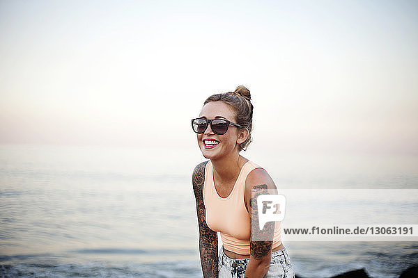 Happy woman in sunglasses standing at beach against sky