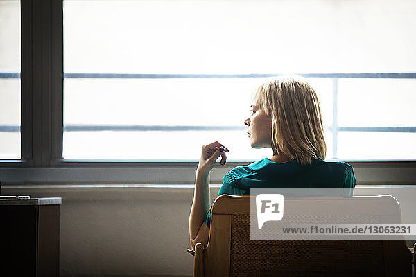 Rückansicht einer Frau  die weg schaut  während sie auf einem Sessel am Fenster sitzt