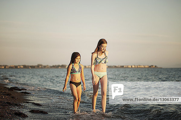 Happy sisters walking in sea against sky during sunset