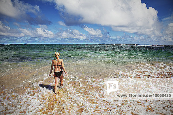 Rear view of woman wearing bikini walking towards sea against cloudy sky