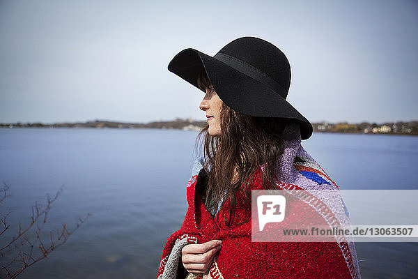 Nachdenkliche Frau mit Hut und Decke am Seeufer stehend