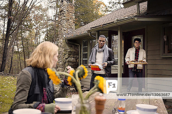 Men bringing food while looking at woman sitting by table outside house