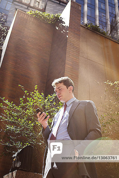Niedrigwinkelansicht eines Geschäftsmannes  der ein Smartphone benutzt  während er an Gebäuden in der Stadt steht