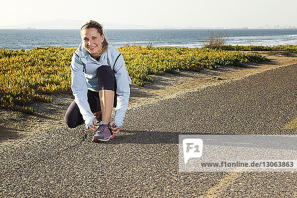 Porträt einer Frau  die Schnürsenkel bindet  während sie auf der Straße gegen das Meer kauert