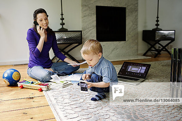 Son playing with technology while mother talking on phone and doing paperwork at home