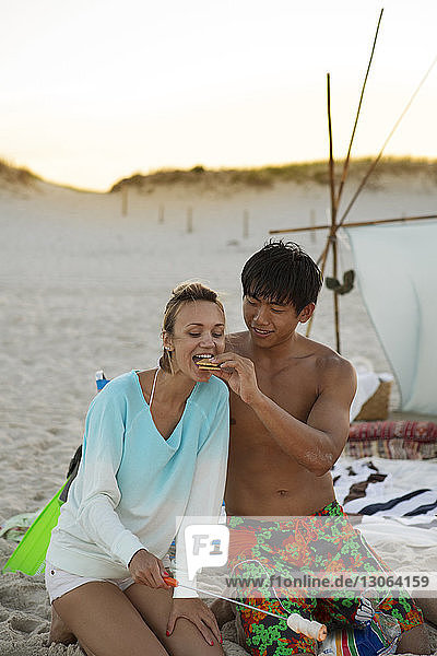 Mann füttert Freundin mit Keksen  während er am Strand am Lagerfeuer kniet
