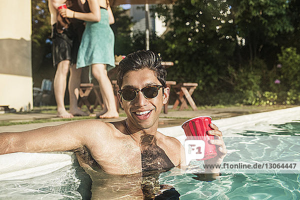 Glücklicher Mann ohne Hemd mit Sonnenbrille  der ein Getränk hält  während er im Pool steht