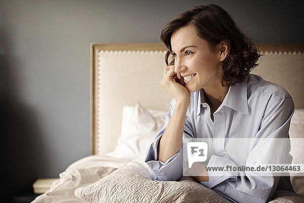 Frau mit Hand am Kinn schaut weg  während sie zu Hause auf dem Bett sitzt