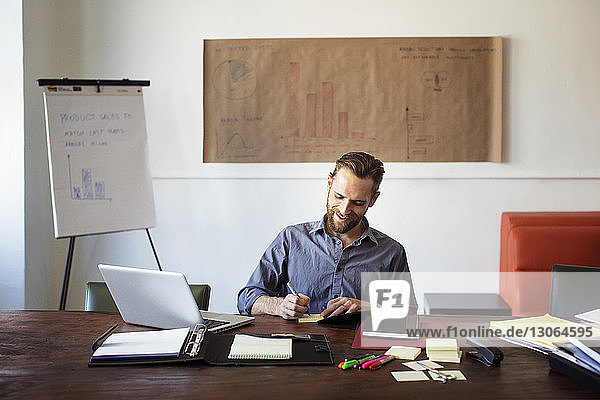 Smiling businessman writing on adhesive note while sitting at desk in office
