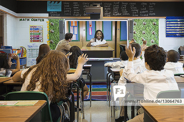 Rear view of students waving hands while looking at photograph on projection screen in classroom