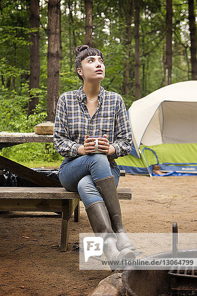 Woman looking up while sitting on picnic table against tent in forest