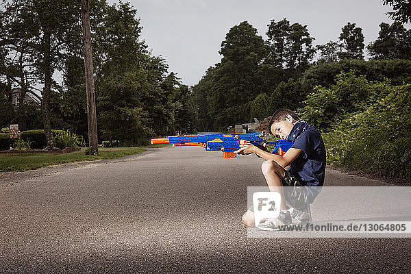 Verspielter Junge zielt mit Spielzeugpistole  während er auf dem Fußweg gegen den Himmel kauert