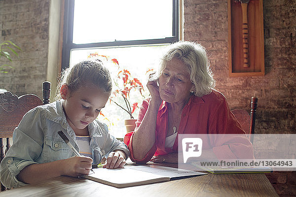 Woman looking at granddaughter doing homework while sitting at table